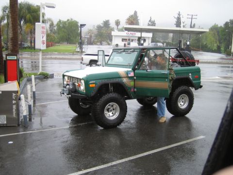 1985 Ford Bronco - A green and white Ford Bronco with gold stripes is parked in a wet lot, with a person standing by the open driver's door.