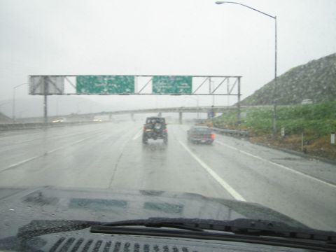 1985 Ford Bronco - A Jeep Wrangler drives on a wet highway under an overcast sky, with another car visible ahead.