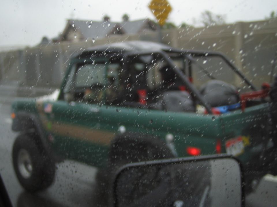 Dark green open-top Ford Bronco with roll cage, seen from rear through rainy glass