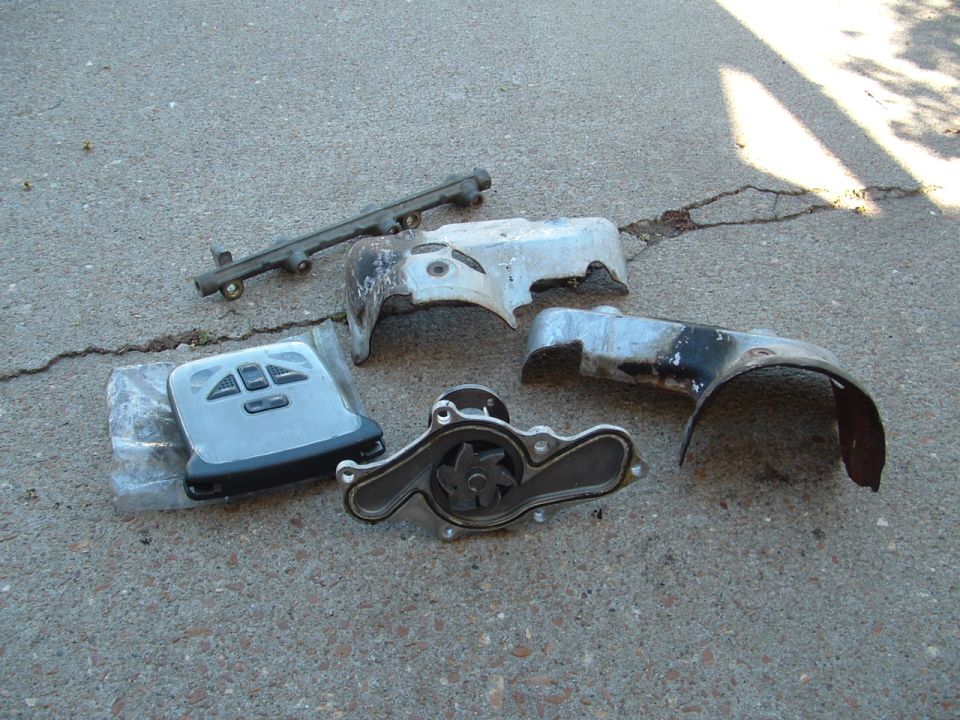 Water pump, timing covers, fuel rail and a window switch panel laid out on cracked concrete