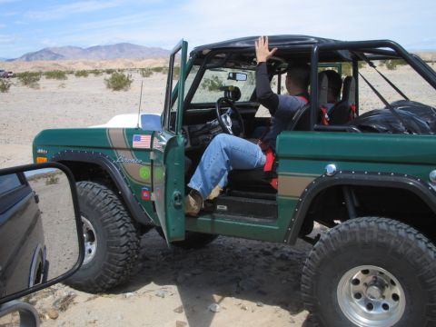 1985 Ford Bronco - A green 1978 Ford Bronco is parked in a desert landscape with its driver's door open and a person waving.