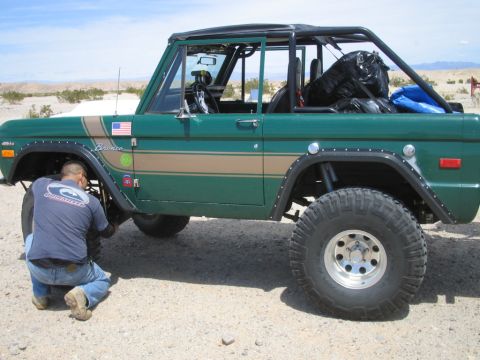 1985 Ford Bronco - A green 1977 Ford Bronco with a tan stripe and large off-road tires is being worked on by a person in a desert environment.