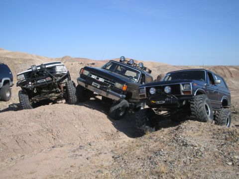 1985 Ford Bronco - Several heavily modified SUVs tackle a steep dirt incline in a desert landscape.