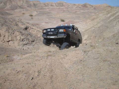 1985 Ford Bronco - A lifted Ford Bronco navigates a steep, sandy incline in a desert landscape.