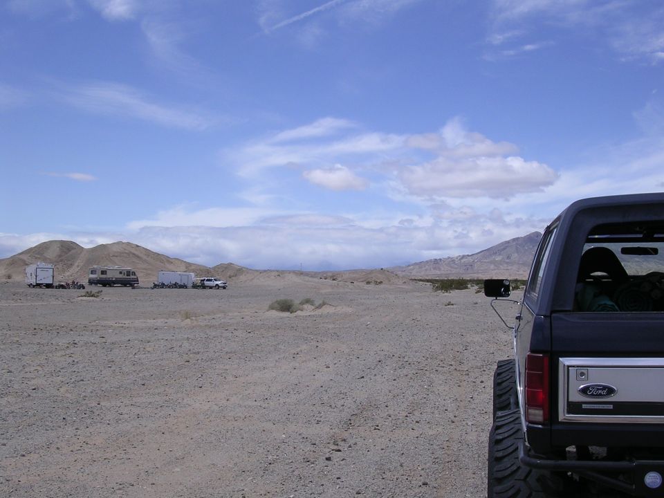 Rear of dark Ford SUV on off-road tires in desert with RV and hills behind