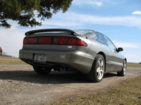 1996 Ford Probe - Low 3/4-rear view of a gunmetal-gray Ford Probe GT hatchback parked on a gravel drive under an evergreen tree, showing the full-width rear…