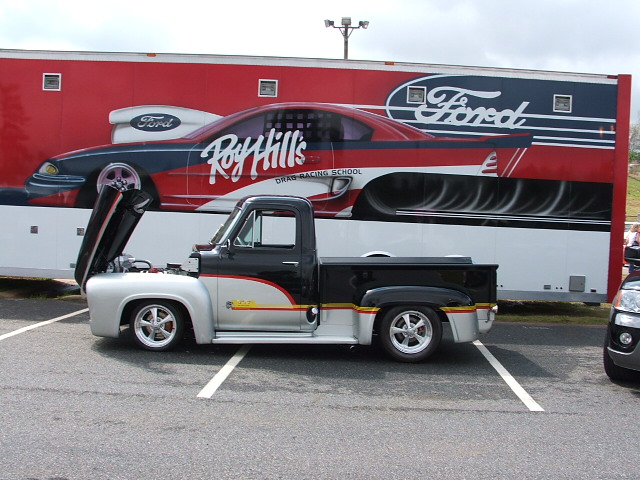 1956 Ford pickup truck, two-tone black and silver, hood open, parked in front of a trailer with a drag car graphic. Another vehicle partially visible on right