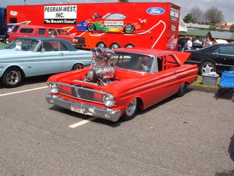 1966 Ford Ranchero - A heavily modified red pickup truck with an exposed engine sits parked next to a light blue classic car, with a large red trailer in the ba…