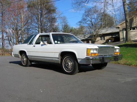 1985 Ford LTD Crown Victoria - A white sedan is parked on a street with a house and trees in the background.