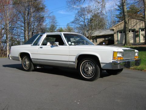 1985 Ford LTD Crown Victoria - A white Ford sedan is parked on a residential street with a house and trees in the background.