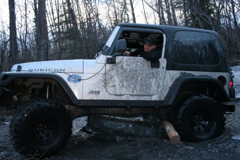 1995 Jeep Cherokee - A driver steers a muddy Jeep Rubicon through a challenging off-road course in a wooded area.