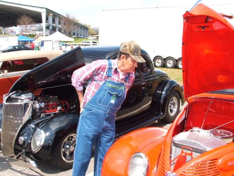1966 Ford Ranchero - Man inspects engine bay of a black classic car at an outdoor car show.