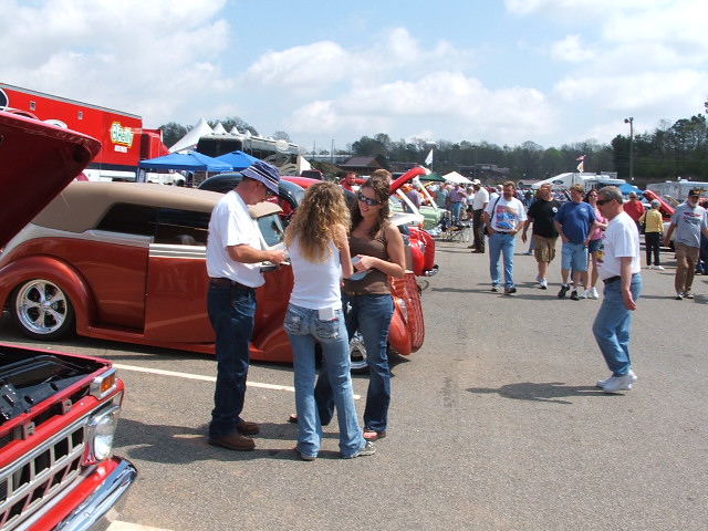 Orange custom convertible hot rod with tan top, people talking near it. Red truck in foreground