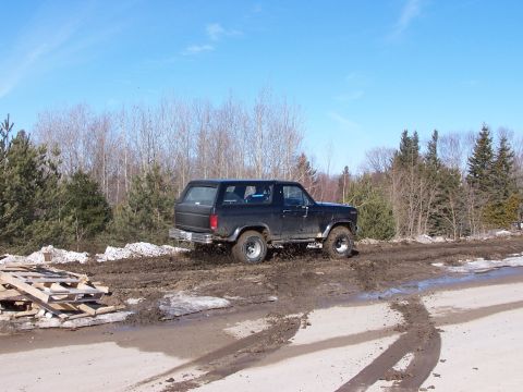 1986 Ford Bronco - This SUV is navigating a rough, muddy terrain with a wooded area behind it.