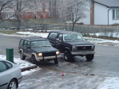 1986 Ford Bronco - Two SUVs, a Ford Bronco and a Jeep Cherokee, are parked on a street with snow on the ground.