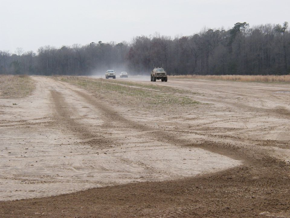 Three vehicles driving on a dusty dirt road with trees in the background