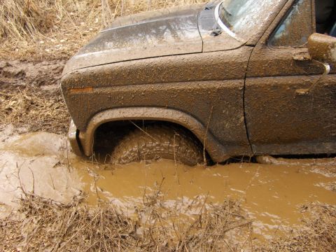 1986 Ford Bronco - A heavily mud-caked vehicle is stuck in a muddy mire, with its front tire deep in the brown water.
