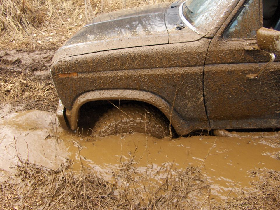 Mud-covered 1980s era truck or SUV stuck in a muddy puddle, front wheel submerged