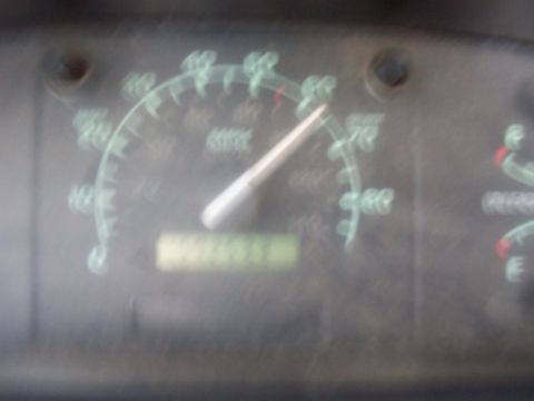 1994 Ford F-150 - Blurry close-up of a dusty Ford truck speedometer face with dual MPH/km-h scales and a mechanical odometer strip below the needle, too out…