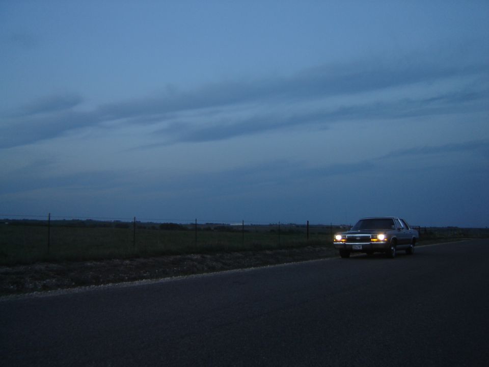1988 Ford Crown Victoria LTD driving on a road at dusk, headlights on
