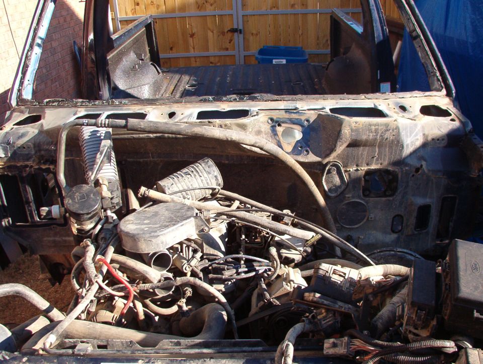 Dusty engine bay of a stripped truck with wiring and hoses, outdoors by a wood fence