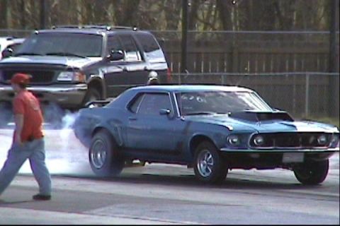 1969 Ford Mustang - A blue 1969 Ford Mustang performs a burnout on the drag strip, with an SUV in the background.