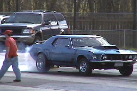 1969 Ford Mustang - A blue 1969 Ford Mustang performs a burnout on a drag strip, with an SUV in the background.