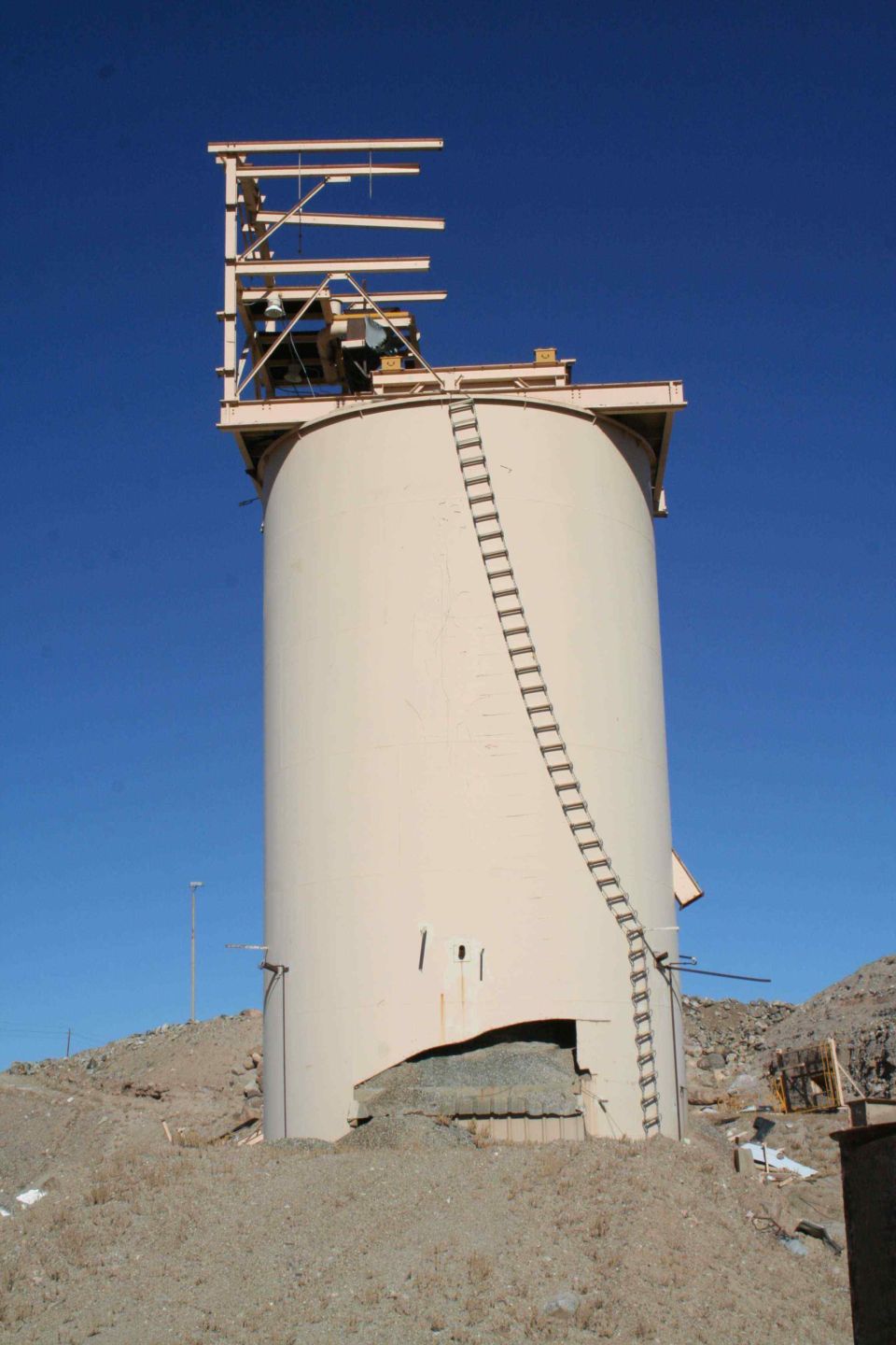 Large industrial silo with a ladder on the side, against a clear blue sky. Some damage is visible at the base