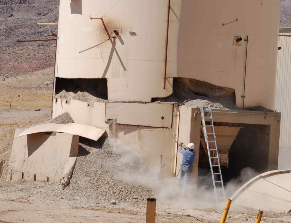 Worker on ladder at damaged industrial silo, dust rising. Supermotors.net logo visible