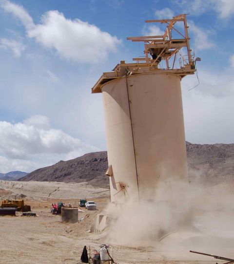 1979 Ford Bronco - Dust billows from the base of a large industrial silo, with several vehicles and construction equipment visible in the arid landscape.