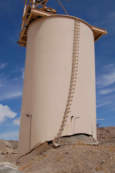 1979 Ford Bronco - A large, beige industrial silo appears damaged at its base, with a ladder running up its side under a clear blue sky.