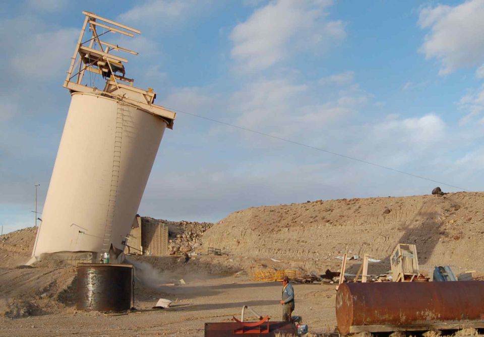 Tilted industrial silo with scaffolding against a blue sky, dusty ground, and a man standing nearby