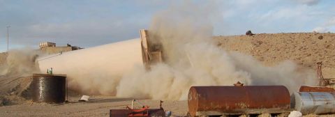 1979 Ford Bronco - A large structure collapses, kicking up a massive dust cloud at an industrial site. Rusty tanks are in the foreground.