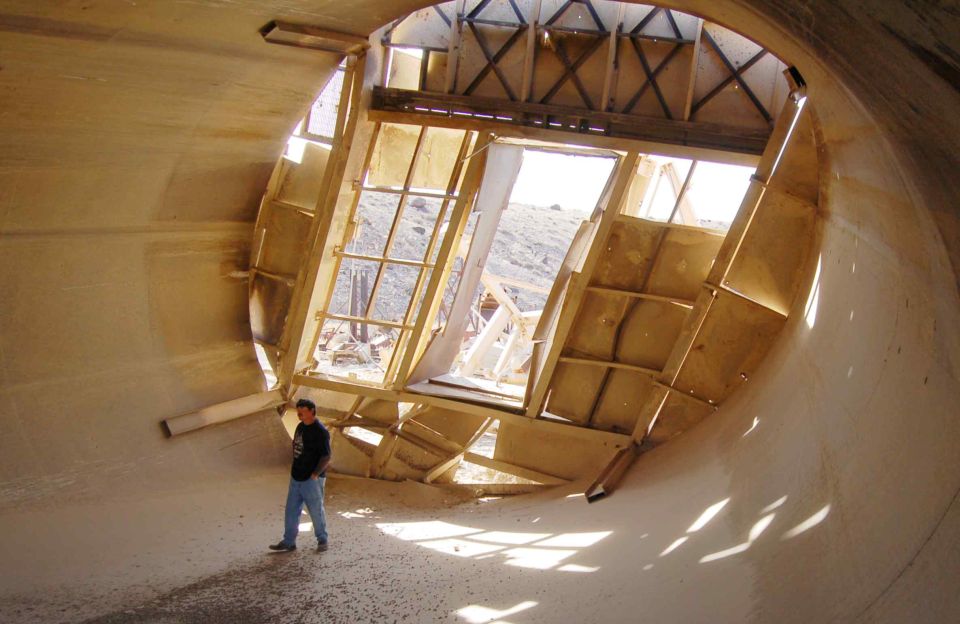 Man in jeans and dark shirt inside a large, unfinished concrete tunnel with wooden framing and windows, casting shadows
