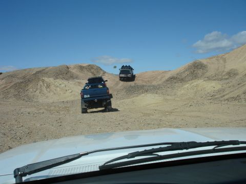 1996 Ford Bronco - Two blue trucks navigate a dusty desert terrain with rolling hills and a bright blue sky overhead.