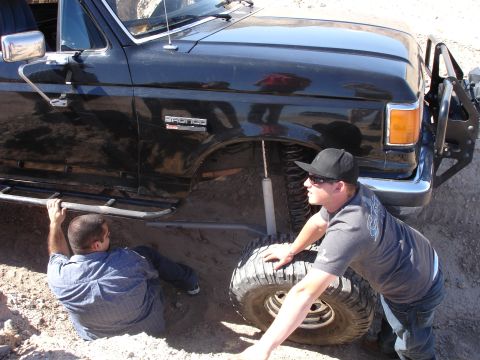 1996 Ford Bronco - Two men work on a lifted black Ford Bronco stuck in the dirt with large off-road tires.
