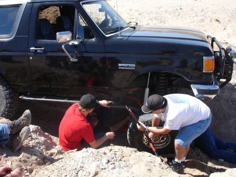 1996 Ford Bronco - People work on the front passenger wheel of a black 1996 Ford Bronco stuck in the dirt.