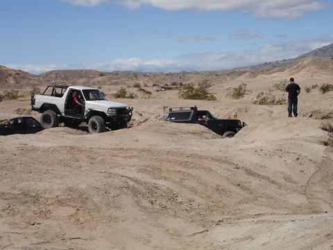 1996 Ford Bronco - Two off-road vehicles navigate challenging desert terrain with one SUV partially submerged in sand.
