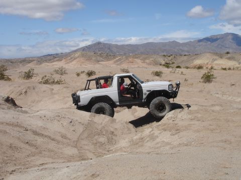 1996 Ford Bronco - A white off-road SUV navigates a sandy desert landscape under a partly cloudy sky.