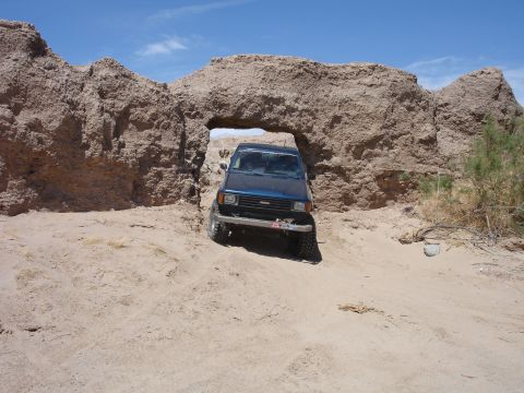 1996 Ford Bronco - A blue SUV navigates through a natural rock archway in a sandy, desert environment under a clear blue sky.