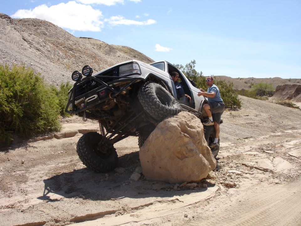 White caged Ford Bronco flexed over a desert boulder with two spotters, winch bumper and lights