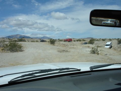 1996 Ford Bronco - Multiple vehicles navigate a dusty desert terrain under a cloudy sky, seen from the perspective of a driver.