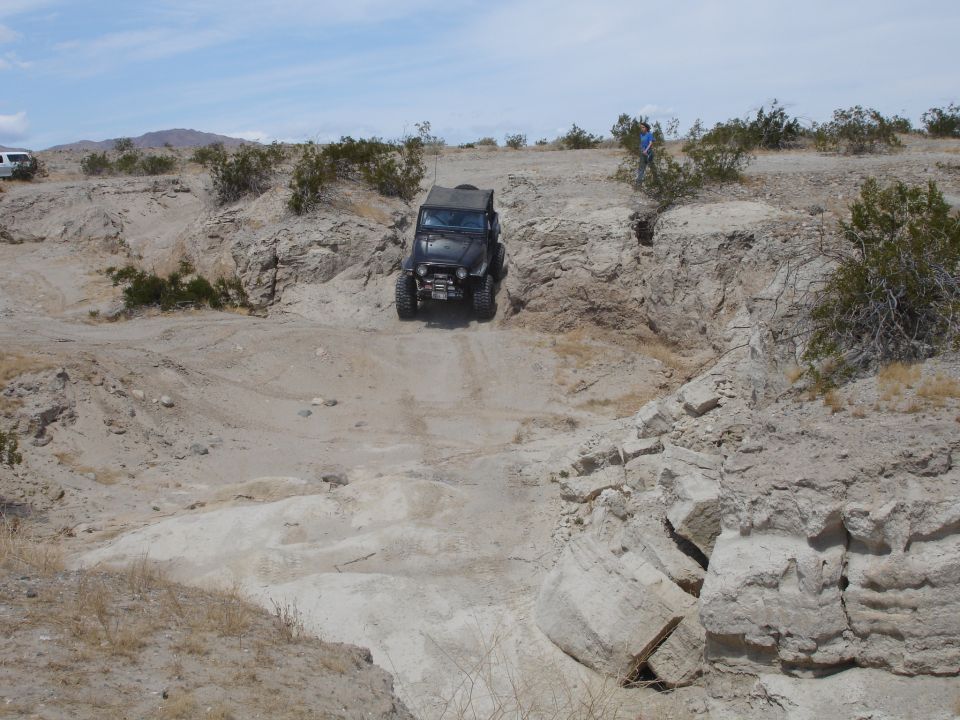 Jeep Wrangler driving down a rocky, off-road trail in a desert landscape. Another white SUV is visible in the distance