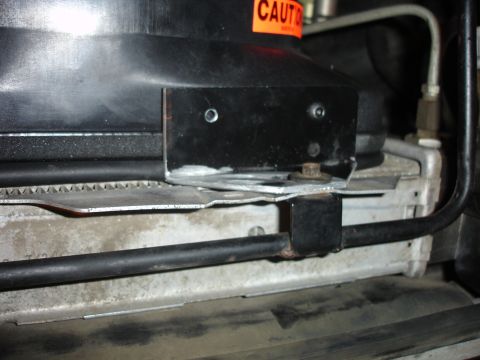 1996 Ford Bronco - Tight close-up under a truck's hood at the radiator core support, showing a bolted-on black steel bracket, a bent aluminum shroud/heat shie…