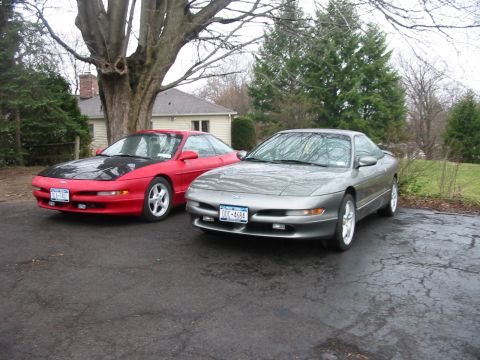 1996 Ford Probe - Two Ford Probes, a red one and a silver one, are parked on a wet driveway in front of a house.