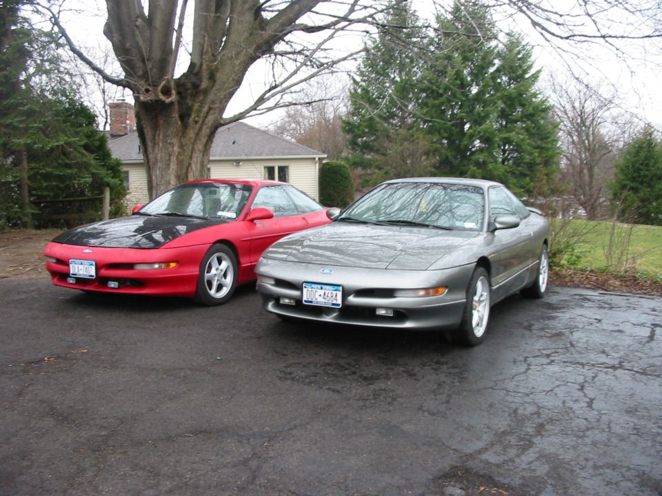 Two Ford Probes, one red with black hood, one silver, parked on a wet driveway