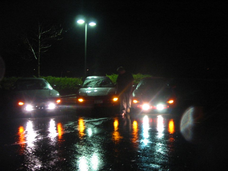 Three cars parked at night, headlights on, with reflections on wet pavement