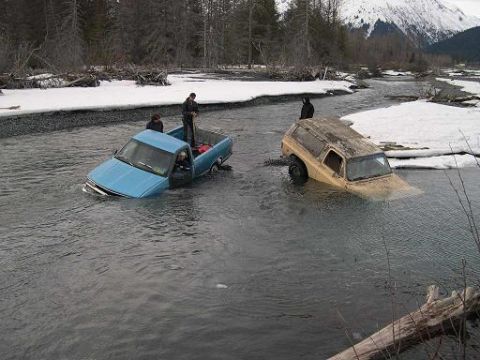1990 Chevrolet Suburban - Two SUVs are stuck in a river with snow on the banks.