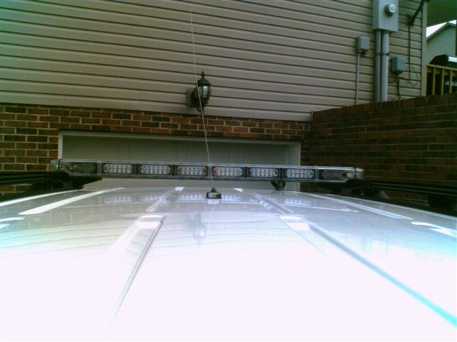 Silver SUV roof with low-profile LED light bar and whip antenna mounted, brick wall behind