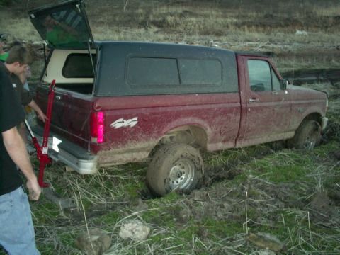 1994 Ford Bronco - A red pickup truck is stuck in the mud with its rear tire submerged.